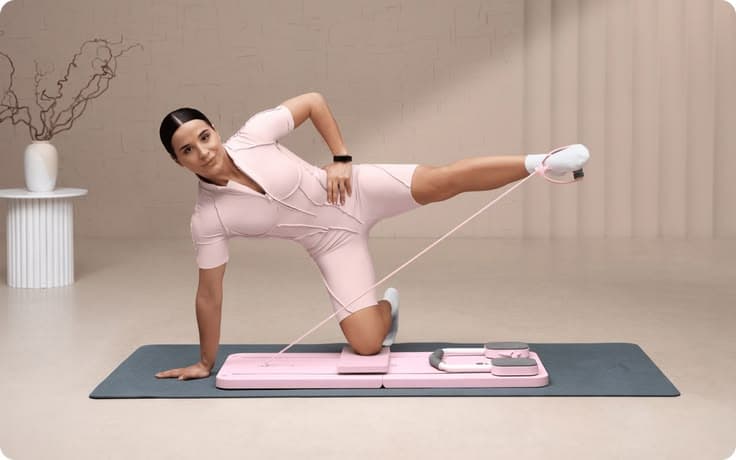 A woman performs a side leg lift on the Pilatease board in a sunlit studio.